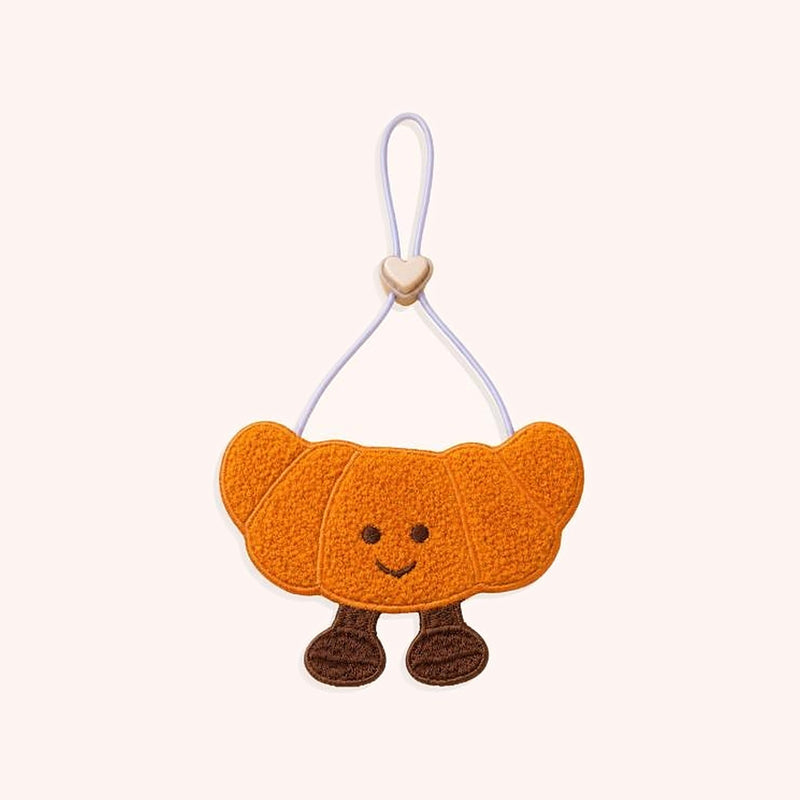 Product photo: Little Croissant Pet Bib — A croissant-shaped plush bib with a smiling face and little feet, on a soft adjustable cord.